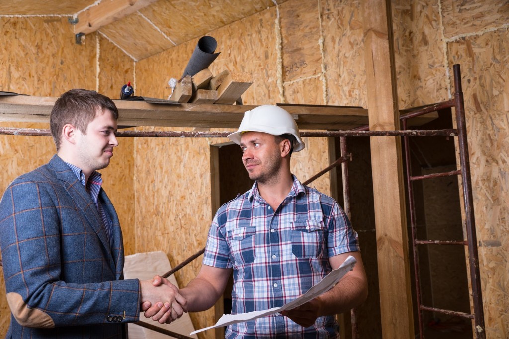 Young Male Architect and Construction Worker Foreman with Building Plans Shaking Hands Inside Unfinished House with Exposed Particle Plywood Boards